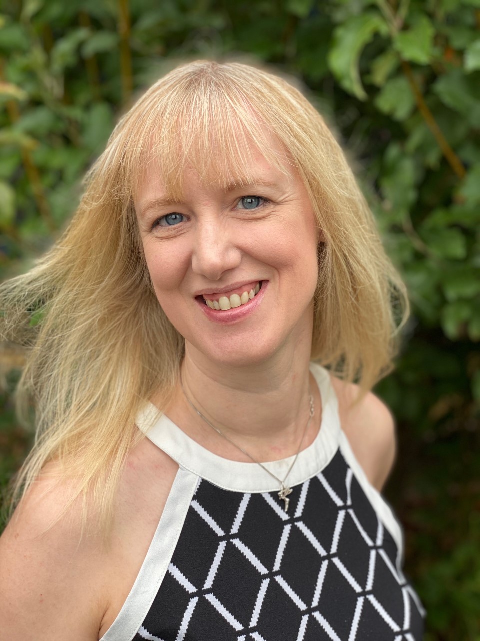 Jeanette Stampone, children's author. This photo is a head and shoulders shot. Jeanette has long straight blonde hair with a fringe, blue eyes and pale skin. She is smiling at the viewer. She is wearing a black and white patterned top, sleeveless. She's standing in front of a leafy green shrub.