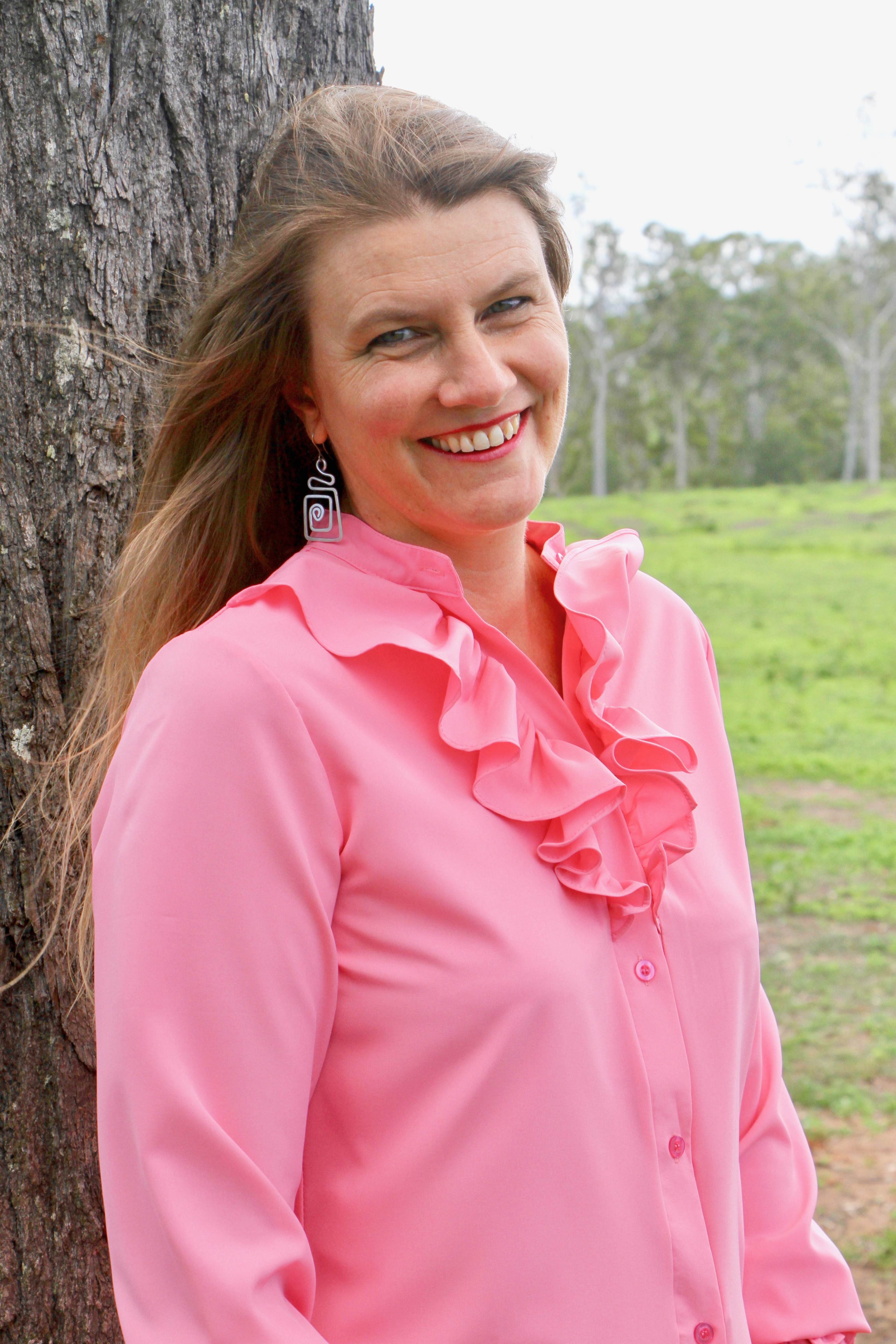 Author Kathryn Apel stands against a tree trunk. She has long blonde hair, is wearing dangling silver earrings and a bright pink shirt with a ruffled collar.