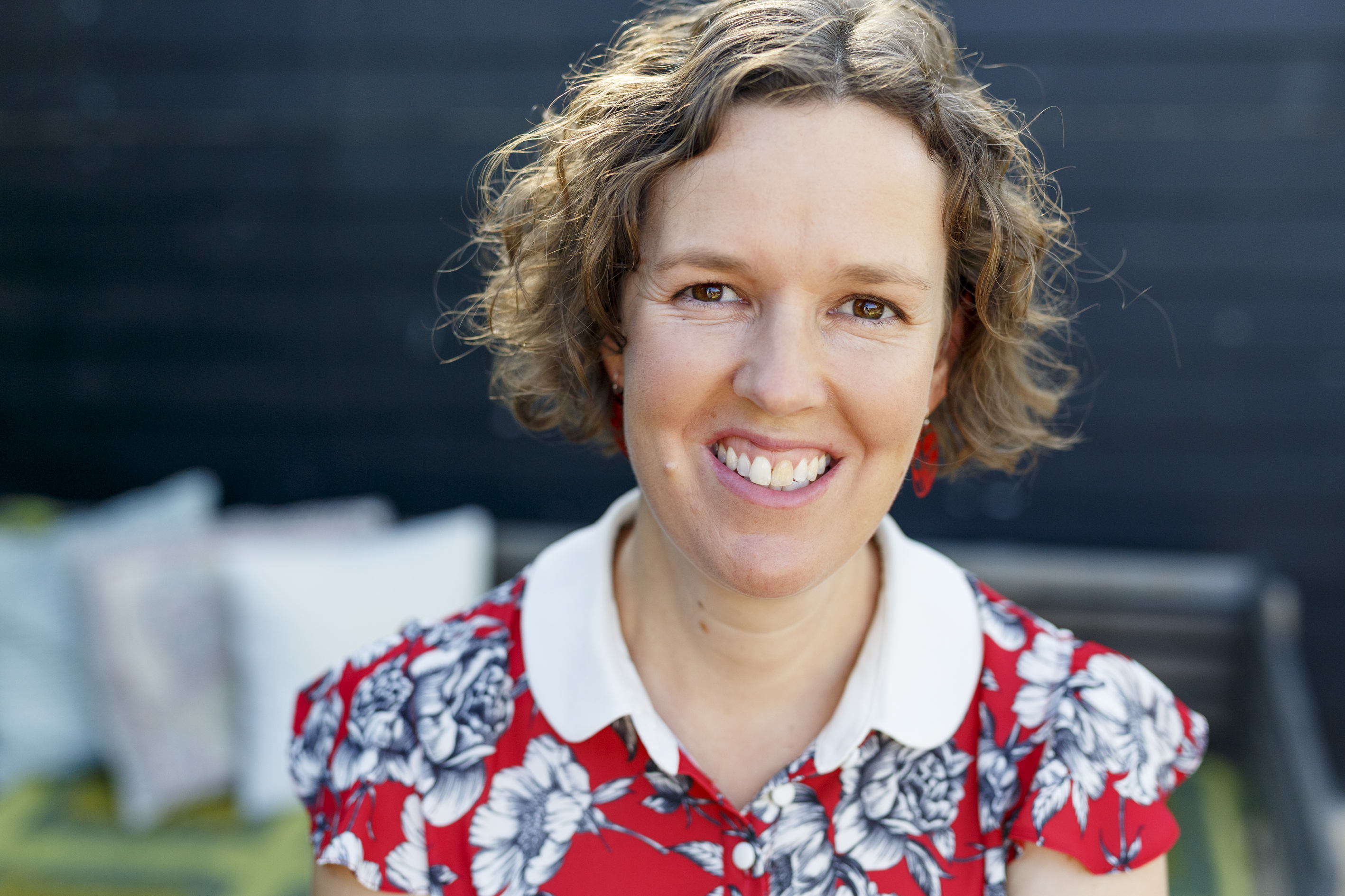The image shows a portrait photo of children's writer HM Waugh. She has fair skin, brown eyes, short light-brown curly hair. She's wearing a red dress with a black and white floral pattern and she wears red earrings. She's smiling at the viewer..
