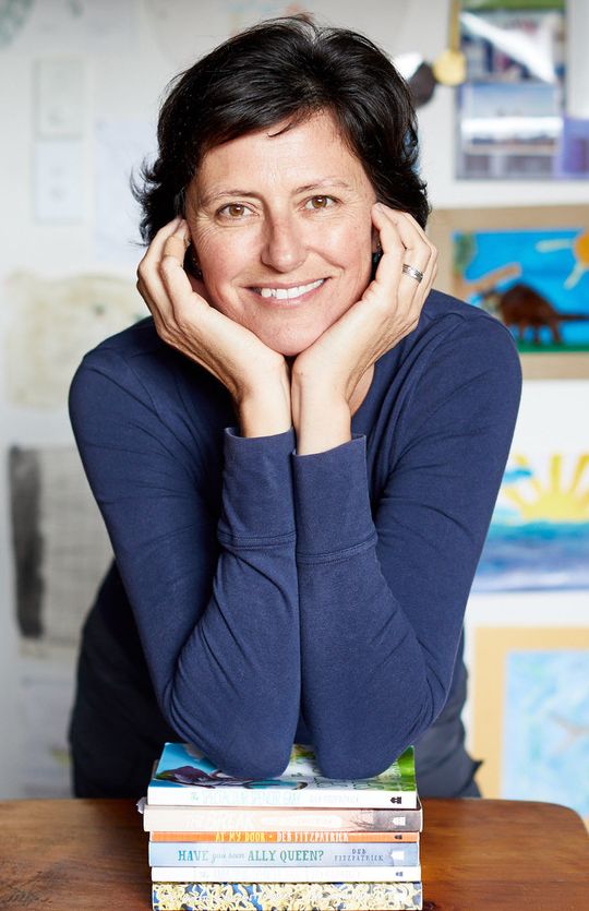 Photo shows author, Deb Fitzpatrick, leaning on her elbows on a stack of books. Deb has pale skin and short dark hair. She's wearing a long-sleeved dark blue shirt and is smiling at the viewer.