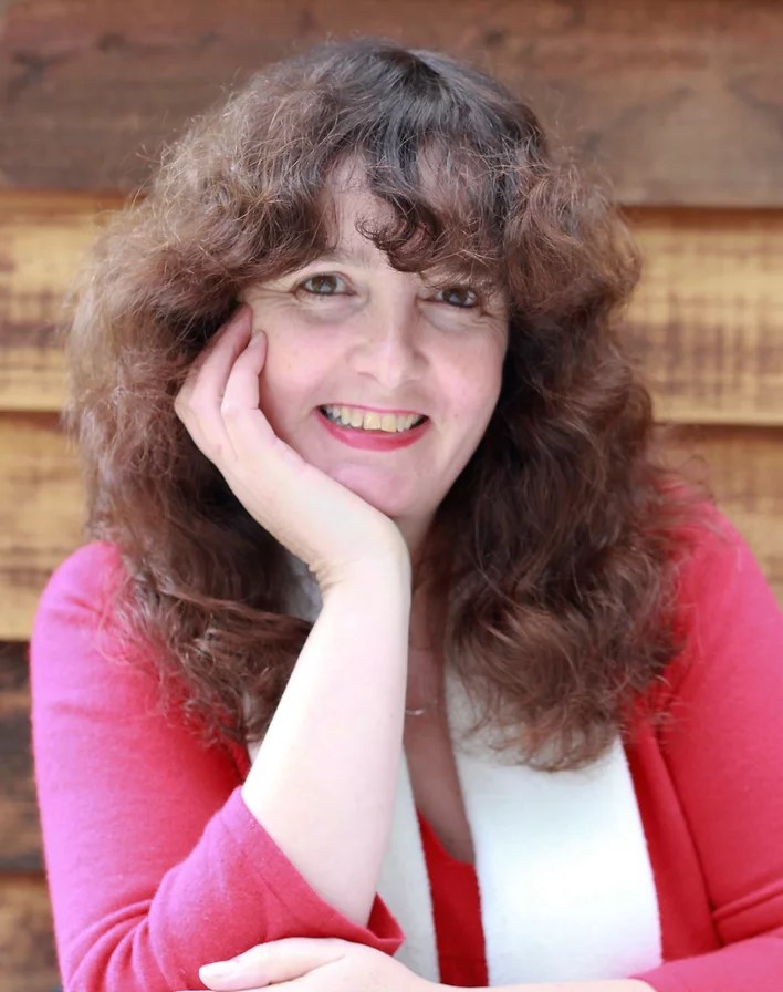Image shows a head-and-shoulders photo of author Dee White. Dee has pale skin, brown eyes and shoulder length wavy brown hair. She is resting her chin on her right hand and smiling at the camera. She's wearing a bright pink top.