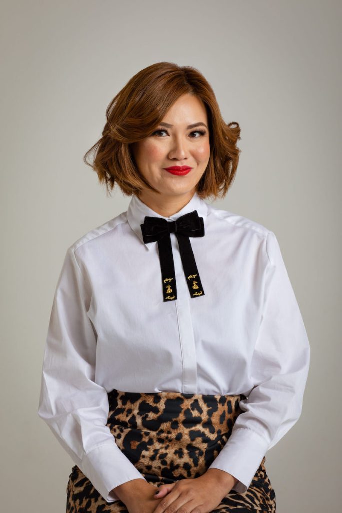 Image shows a photo of author Shirley Marr, a first generation Chinese-Australian. Shirley has short brown hair and bright red lipstick. She's wearing a white button-through shirt with a black bow feature, and a leopard skin print skirt. She's sitting with her hands in her lap.