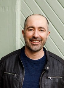 James Foley (author-illustrator). James has pale skin and a dark beard. He's wearing a black jacket over a tshirt and standing in front of a white wooden door.