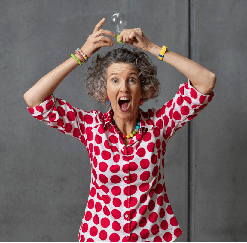A woman in a white shirt with red spots stands holding a light globe over her head with two hands